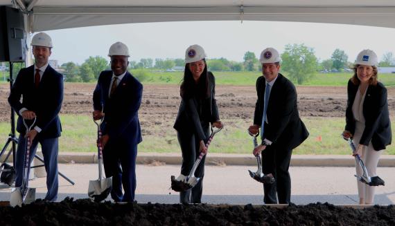 Five people wearing business attire and hard hats hold shovels of dirt under a tent outdoors on a construction site.