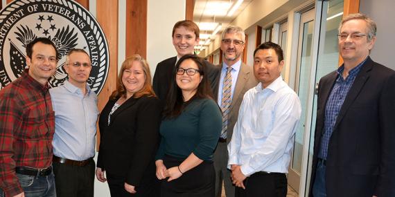 Group of eight smiling people in an office with the Department of Veteran&#039;s Affairs seal behind them