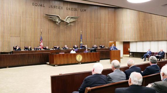 Large wood-paneled and -appointed courtroom with Equal Justice Under Law and large eagle sculpture on back wall, with a panel of people in black robes below it, with court workers, a speaker, and white-haired men in the audience