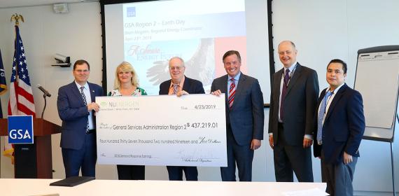Group of six people in business attire next to a GSA-branded podium and a U.S. flag, holding a large check to GSA for $437,219.01