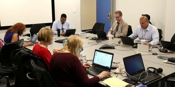 Six people with laptops sitting around a rectangular conference table looking at their screens or each other