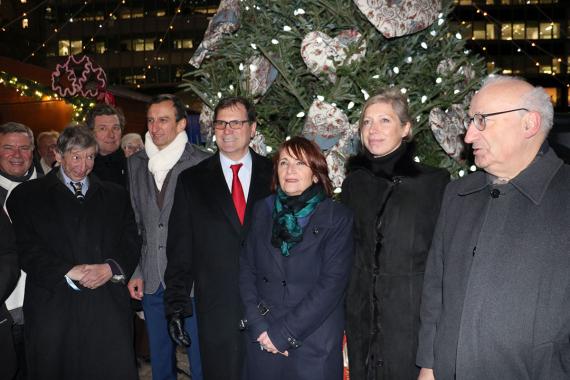 Eight people in winter wear standing in front of an evergreen tree near a holiday-themed market