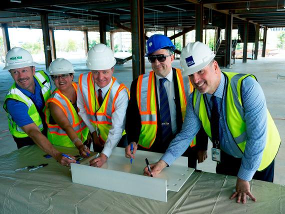 Five smiling people in yellow safety vests and four white hard hats and one blue one stand in an unfinished building and sign a white beam section