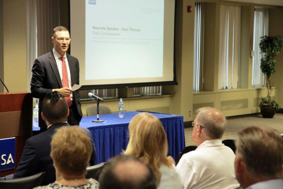 Man in black suit and red tie speaks at a podium to a group of people facing him (photo by Robert Andersen)