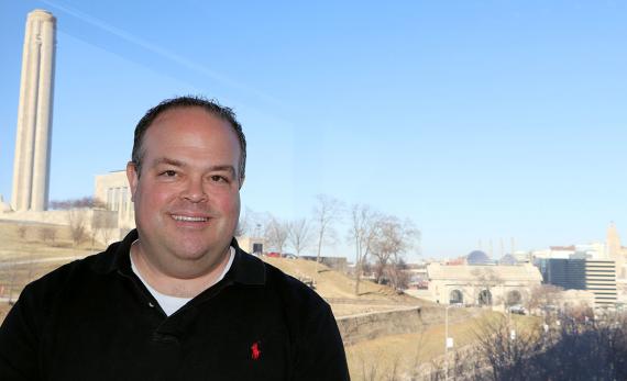 Smiling man in black polo shirt poses in front of a view of a large monument