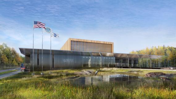 Image of 1-story grey building, with awning to the left. 3 flags fly to the right of the image. A small pond with rocks is to the front side