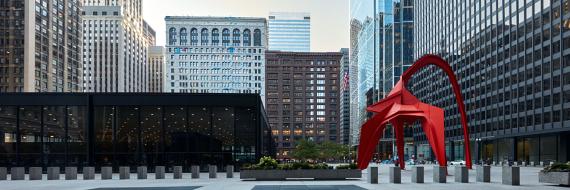 Large red metal sculpture in a plaza in front of black buildings with mirrored windows