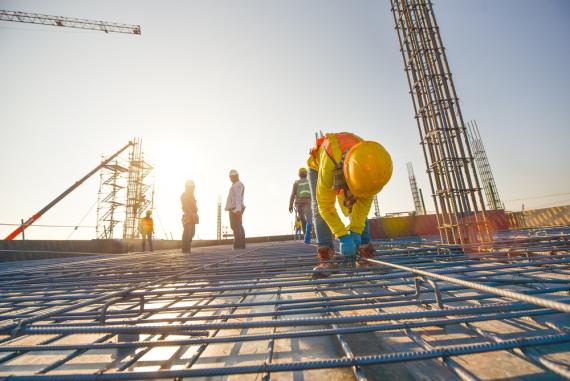 A man in construction attire is bending down working on a mental grid with other construction men in the background