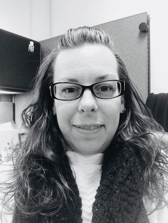 Black and white photo of a person wearing glasses and a scarf sitting at an office desk.