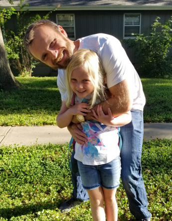 An adult standing in front of a house wearing jeans and a T-shirt and hugging a child.