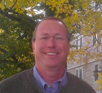 A person with glasses wearing a blue collared shirt and sweater, outside.