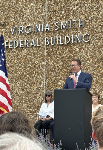 A man with glasses in a blue suit speaks at a podium in front of the Virginia Smith Federal Building&#039;s signage.