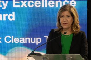 Woman with shoulder length red hair in green shirt and black blazer speaking at podium