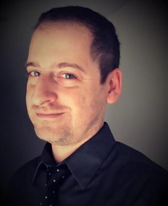A headshot of Ben Baellow, a male with dark hair, wearing a dark collared shirt and polka dot tie. 
