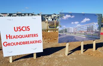 Two signs in dirt in front of a housing development with blue sky above. One sign reads USCIS HQ groundbreaking and the other is a picture of the future building.