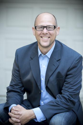 Chris Bolinger, white male with faded hair, sitting, smiling, wearing glasses, a blue business shirt, blue sport jacket and jeans.