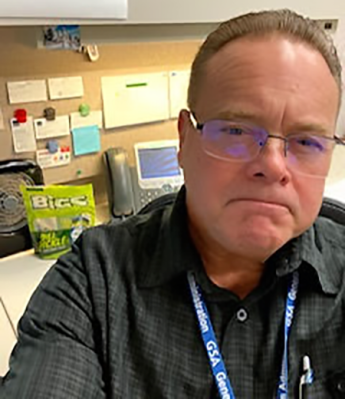 Selfie-style photo of a person with glasses, wearing a business shirt and GSA lanyard, sitting in front of a desk that contains a bag of dill pickle flavored sunflower seeds, a portable fan, and a phone.