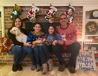 A family of four sits on a fireplace hearth holding two dogs with Christmas and Hannukah decorations around them.