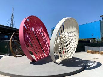 Outdoor sculpture of three large perforated disks, brown, red and white, set at angles that cause interactive shadows, on round cement base with blue sky in background.