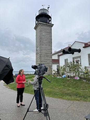 A woman in a red jacket speaking in front of a video camera with a lighthouse in the background