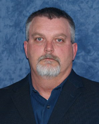 A person with open collared shirt and sport jacket in front of a blue, studio background.