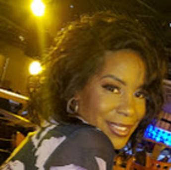 A headshot of Tranita Stanley, a person with brown skin and shoulder length brown hair, head turned, smiling over their shoulder, wearing a black and white blouse. 