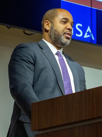 Black man with short cropped hair and beard in gray jacket and purple tie, speaking at a lectern 