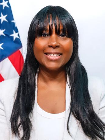 Smiling Black woman with long hair and bangs in a white top and jacket in front of a U.S. flag