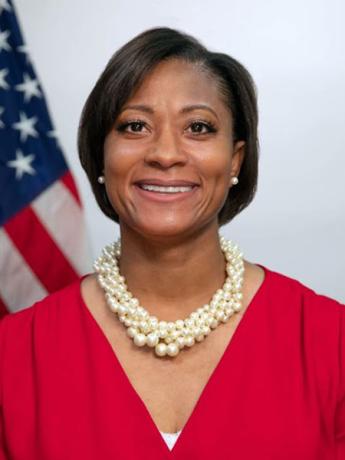 Smiling Black woman with short hair and pearl earrings and necklace, and red top, in front of U.S. flag