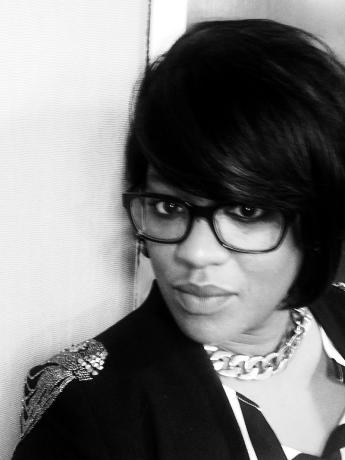 Black and white photo of a Black woman in a bob haircut, dark glasses frames, dark jacket with beaded embellisment and large chain necklace