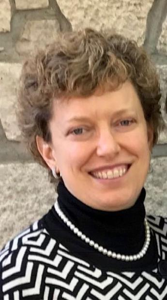 Somewhat blurry headshot of a white woman with short curly hair wearing a black and white turtleneck sweater with a pearl necklace.