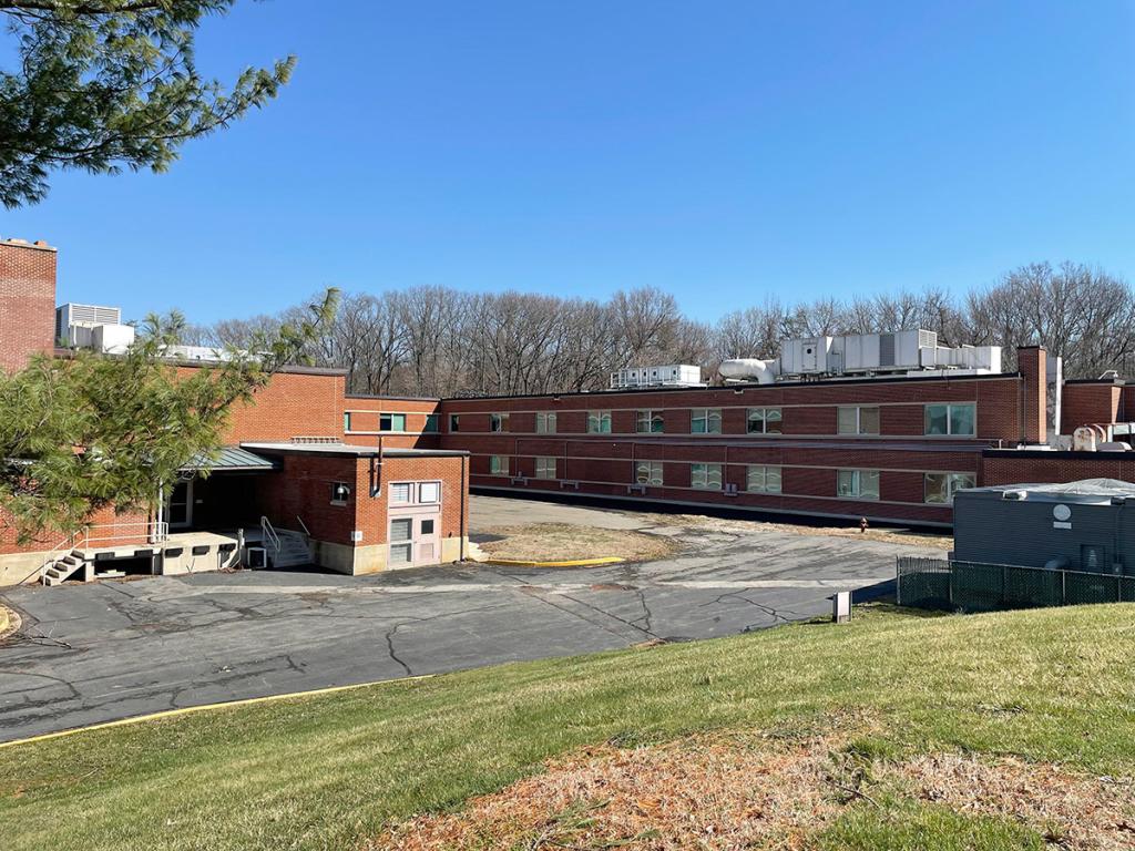 Two-level red brick building with trees behind it, and a driveway and grass in front