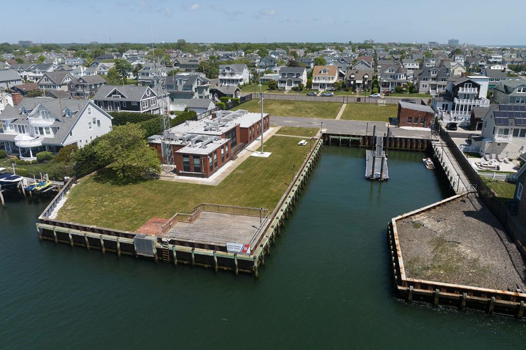 Red brick building with a seawall, boat basin, small dock, deck on a lawn, parking lot and brick maintenance building.