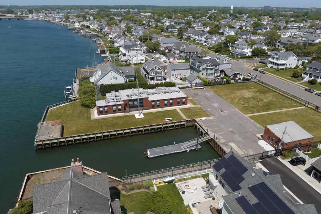 One-story brick building on a river with seawall, deck, boat basin, dock, fence, parking lot, maintenance building. Street, houses nearby.