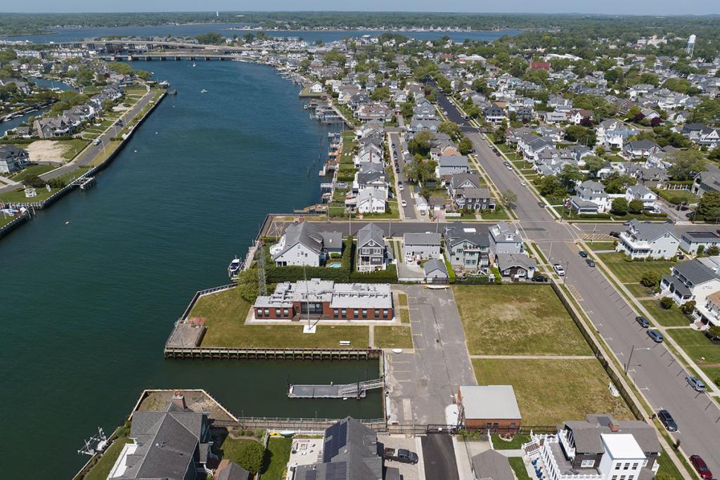 One-story brick building on river with seawall, deck, boat basin, dock, fence, parking lot, maintenance building. Houses on a street.