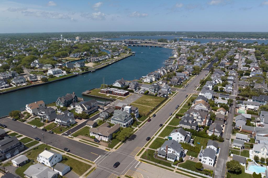 One-story brick building on river with seawall, deck, boat basin, dock, fence, parking lot, maintenance building. Houses on a street.