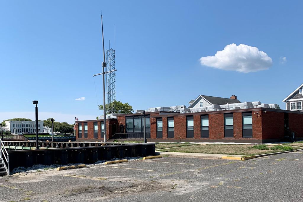 A large communications tower stands near a single-story red-brick building with multiple windows next to a parking lot.