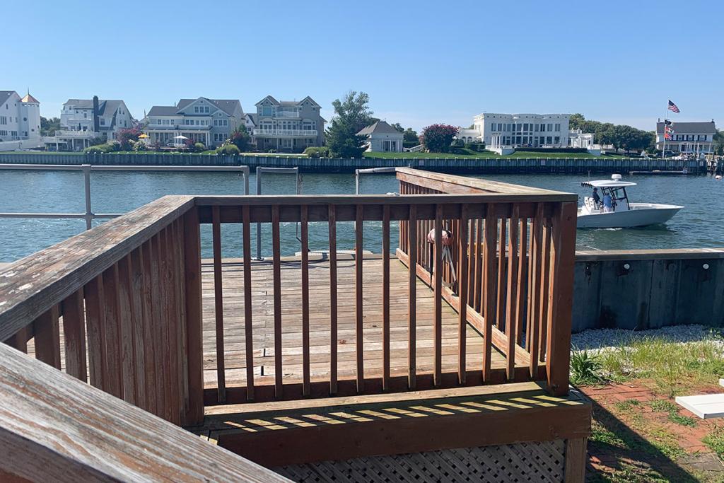 A deck with a wooden rail gives a view of a river with large houses on the opposite bank.