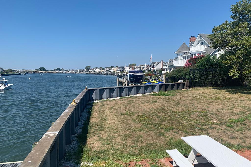 A grass lawn leads to a seawall with a view of a river and houses.