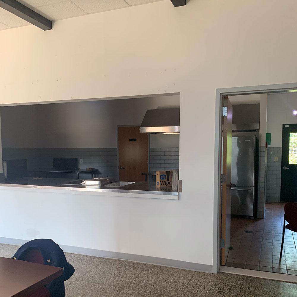 A kitchen with tile floor, refrigerator, exhaust fan, and large pass-through serving window. The walls are white and partially tiled.