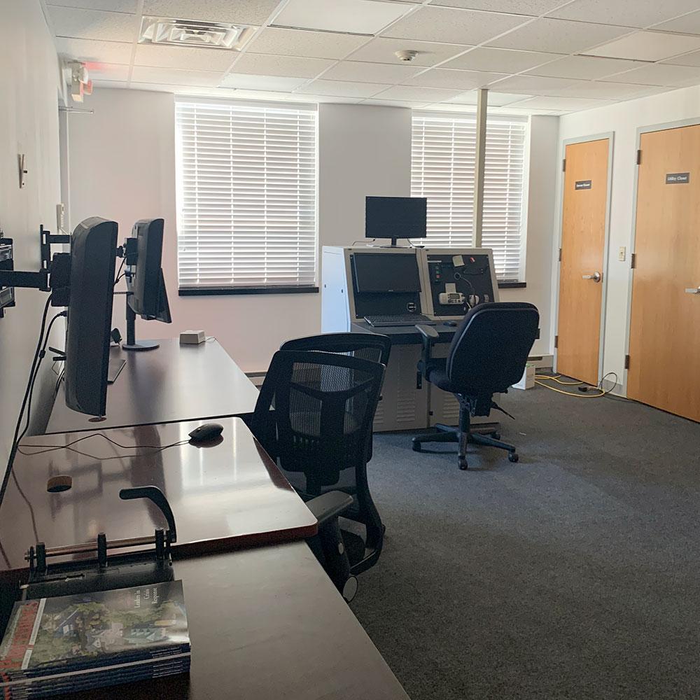 Office with two windows, two doors, desks, two chairs, and control station. White walls and carpeted floors. Blinds on windows.