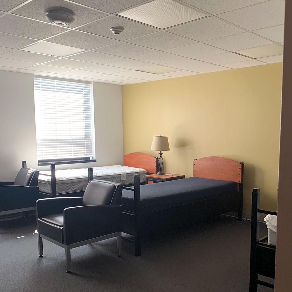 Dormitory room with three single beds, table with lamp, a window with blinds, and two chairs. White and yellow walls and carpeted floors.