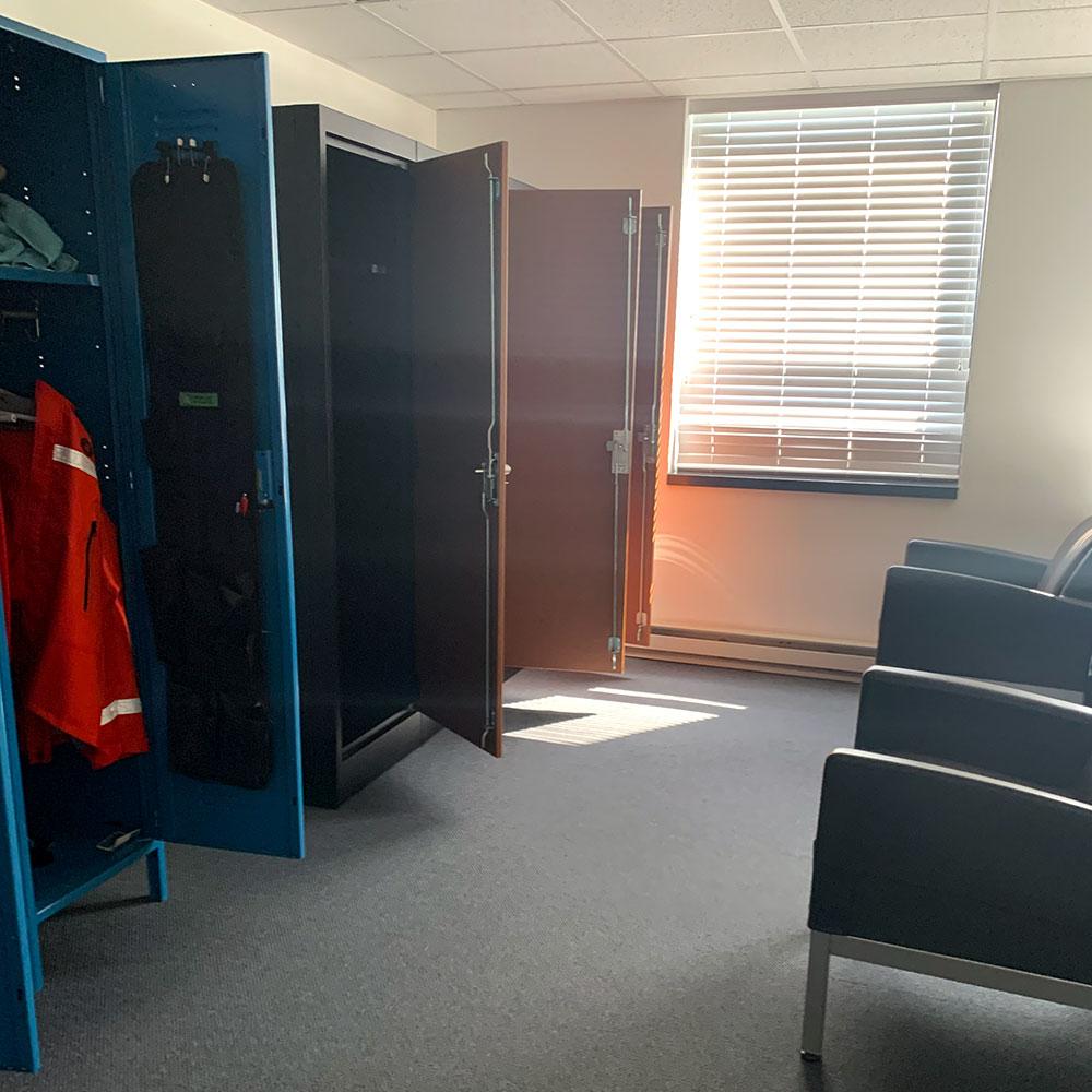 A dormitory room with three open closets, a single window with blinds, and two chairs. The floor is carpeted. The walls are white.