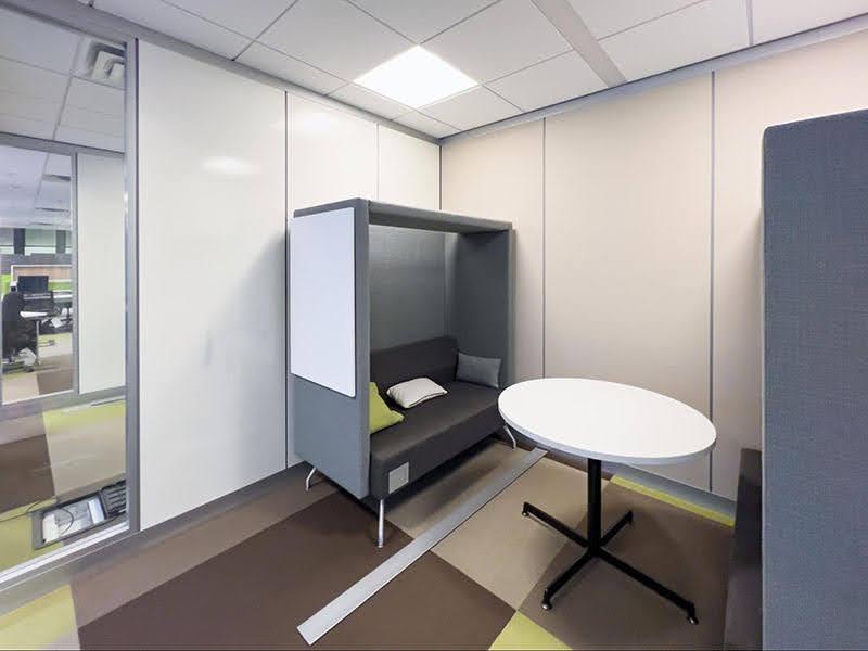 A room with a round, white table and grey cubby or booth and a bench seat among white and glass paneled 