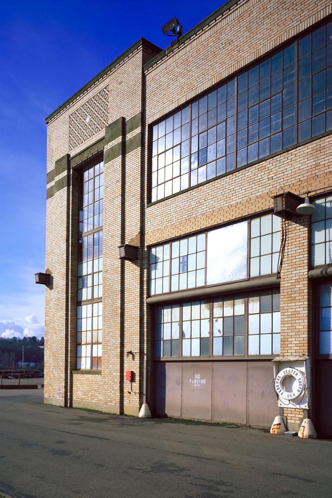 Side of two-level yellow brick building with large, multi-paned windows, and a buoy sign, against a blue sky