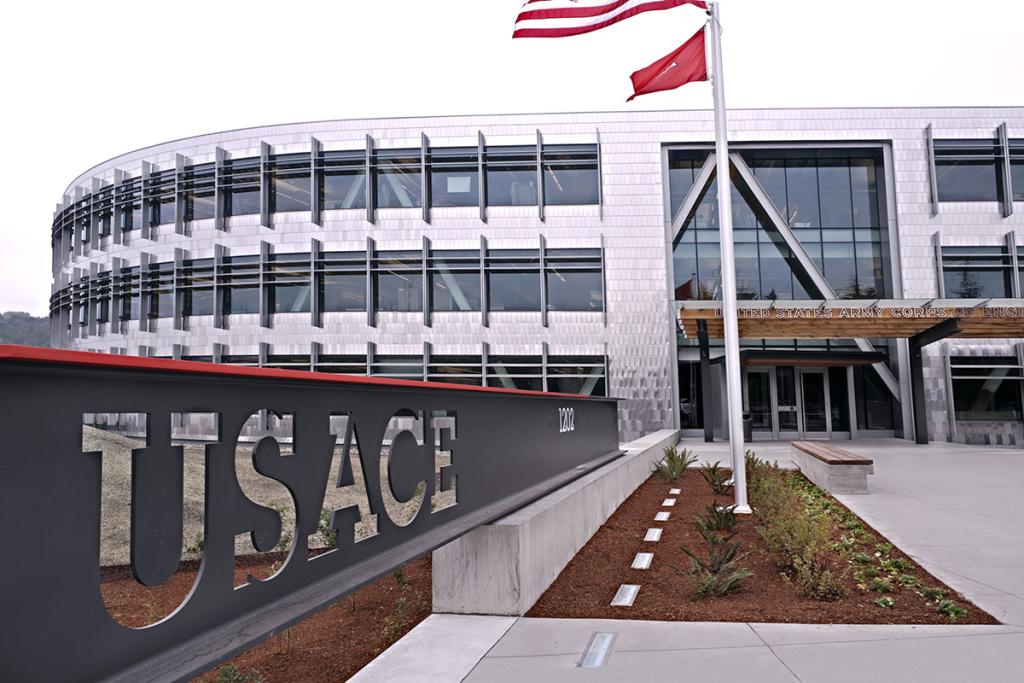 Three level curved silver faceted building facade with partial view of two flags, and USACE sign along walkway to entrance