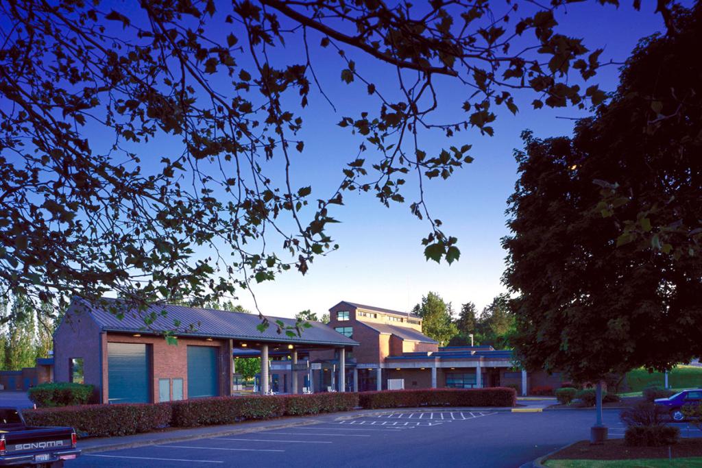 Red brick building complex with low hedges, parking lot and trees, and blue sky at twilight