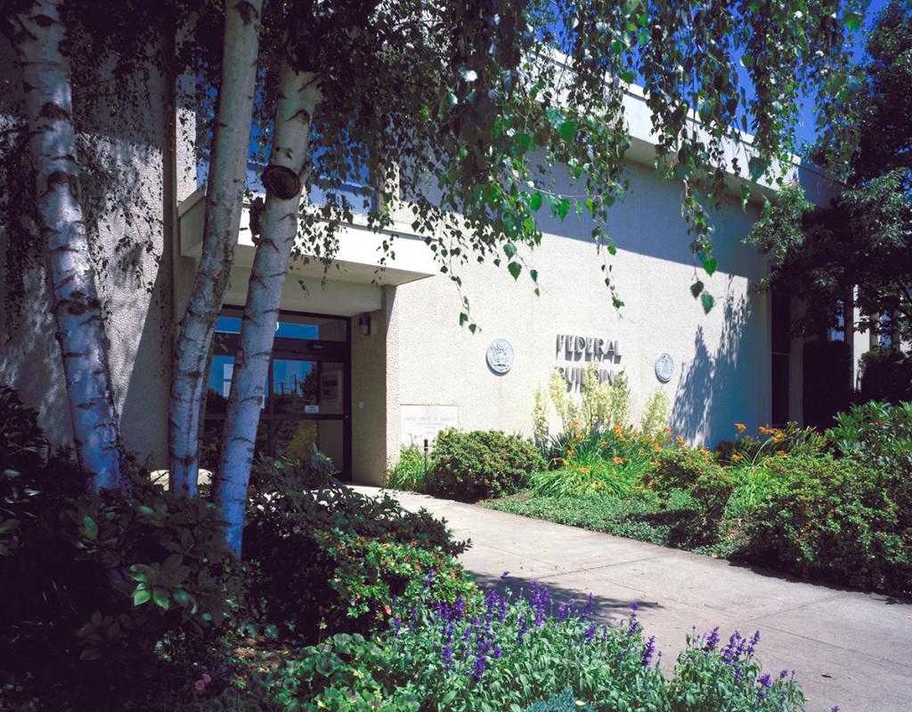 Light stone or stucco building entrance with two seals and lettering Federal Building to the right of the glass door, with trees and flower beds on either side of the walkway