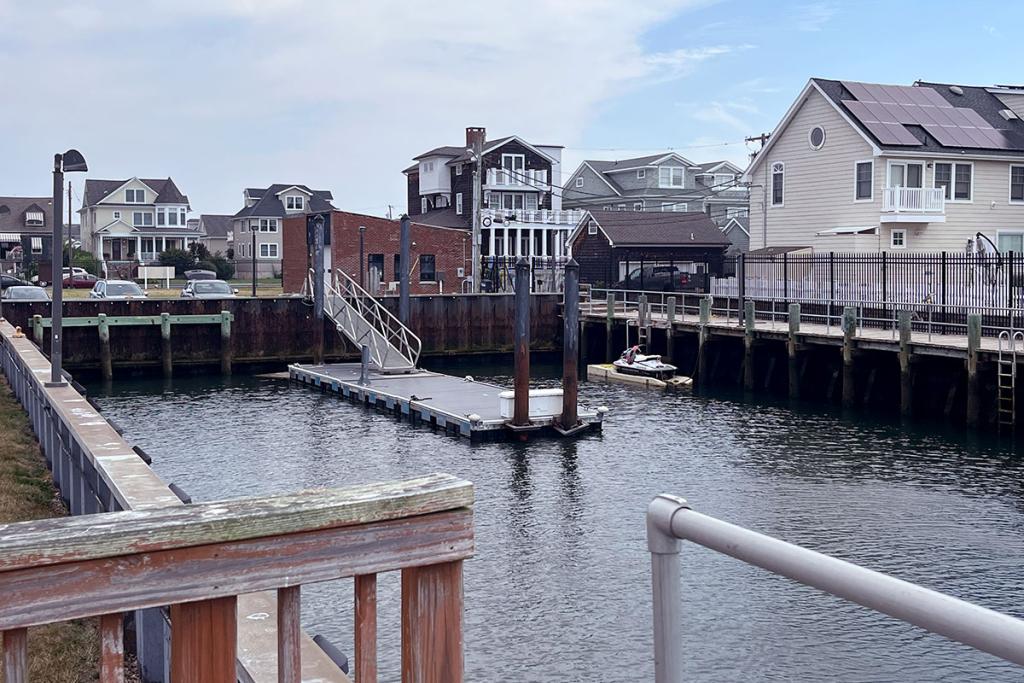 A dock leads to a small boat basin. A small white boat is docked nearby. A fence separates the basin from multiple houses.