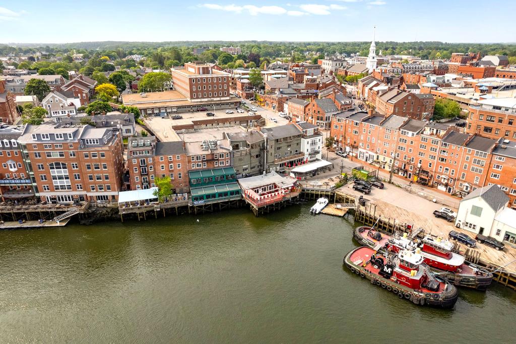 Aerial view of a four-story red brick building with a large one-story annex overlooking a harbor with tugboats and a row of brick buildings and docks on a waterfront.